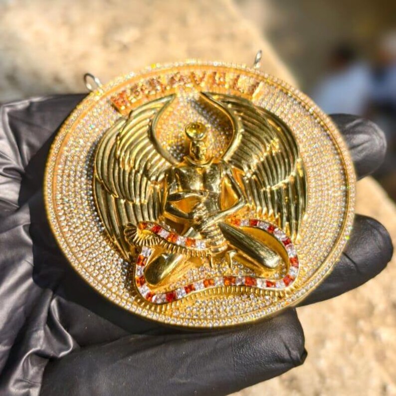 May include: A gold pendant featuring a winged figure with intricate detailing. The pendant is encrusted with numerous small, sparkling stones and has a red and white accent. The word "RENAISSANCE" is visible on the top of the pendant.