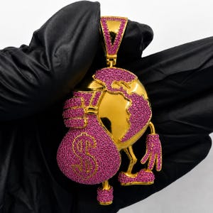 May include: A gold pendant with a globe and money bag design. The globe and bag are covered in pink gemstones. The money bag has a dollar symbol. The pendant has a gold bail.