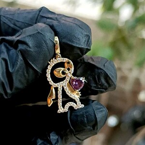 May include: Gold-toned pendant featuring a stylized figure with a heart-shaped purple gemstone. The figure is adorned with small, clear gemstones and has black accents. The pendant is suspended from a gold bail, showcasing a whimsical design.