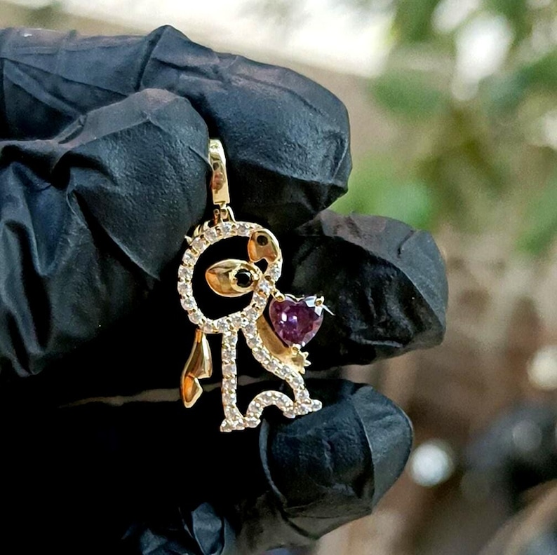May include: Gold-toned pendant featuring a stylized figure with a heart-shaped purple gemstone. The figure is adorned with small, clear gemstones and has black accents. The pendant is suspended from a gold bail, showcasing a whimsical design.