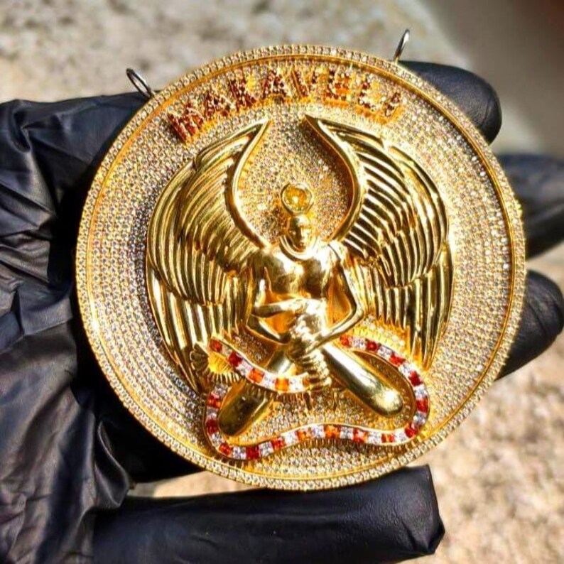 May include: A large, round gold pendant featuring an angel figure with outstretched wings. The pendant is encrusted with small, sparkling stones around the edge and the angel's wings. The word "MAKAEL" is inscribed at the top. A red, white, and orange snake-like design is at the bottom.