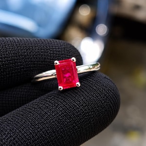 May include: A silver ring featuring a rectangular emerald-cut vibrant red gemstone held in a four-prong setting. The band is smooth and polished, showcasing a clean metal finish that complements the bright, saturated color of the central stone.