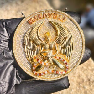 May include: A gold-colored pendant with a detailed design. The pendant features a central figure with outstretched wings, surrounded by a circle of sparkling stones. The word "MAKAVELI" is at the top.