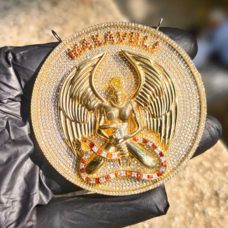 May include: A gold-colored pendant with a detailed design. The pendant features a central figure with outstretched wings, surrounded by a circle of sparkling stones. The word "MAKAVELI" is at the top.