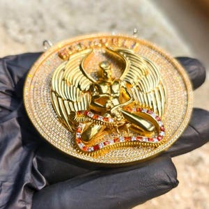 May include: A gold-colored pendant featuring an angel with outstretched wings, detailed with small, sparkling stones. The angel is seated, with a red and white stone accent around the base. The pendant has a circular shape.
