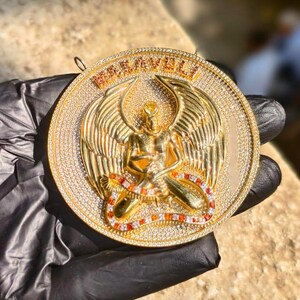 May include: A gold-colored pendant with a detailed design featuring an angel figure with outstretched wings, a snake, and the word "MARVELI". The pendant is encrusted with numerous small, clear gemstones.