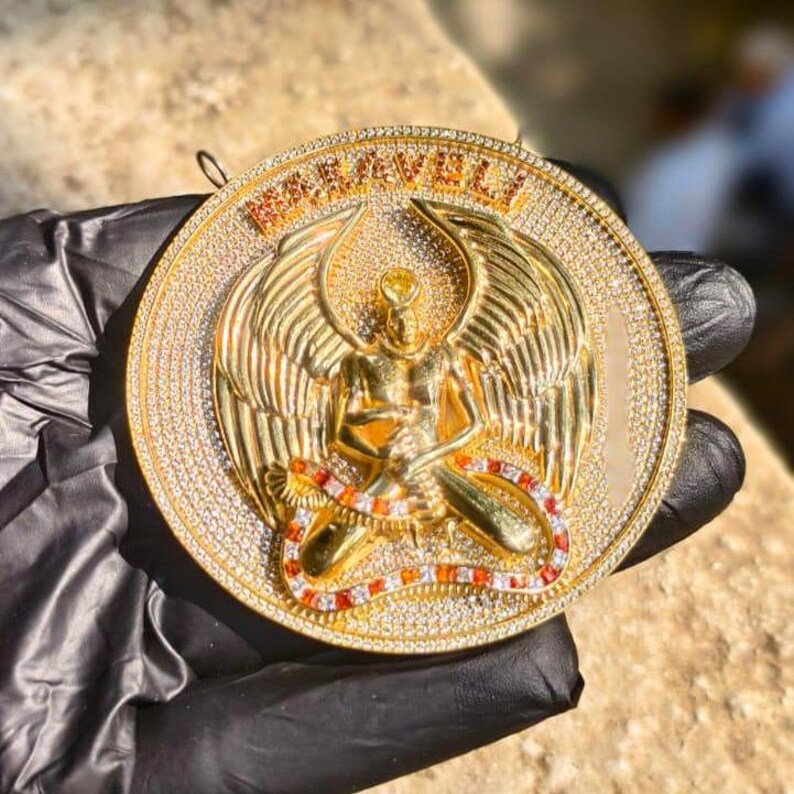 May include: A gold-colored pendant with a detailed design featuring an angel figure with outstretched wings, a snake, and the word "MARVELI". The pendant is encrusted with numerous small, clear gemstones.