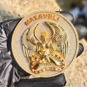 May include: A gold-colored pendant with the word "MARAVELLI" in orange letters. The pendant features a detailed angel figure with outstretched wings, surrounded by small, sparkling stones. The angel is seated with a snake-like design around its legs.