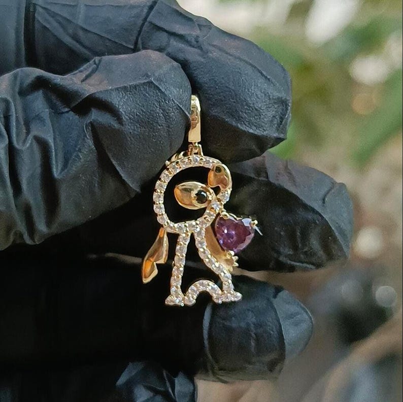 May include: A gold-colored pendant in the shape of a cartoon character holding a purple heart. The pendant is encrusted with small, clear gemstones and features black accents. The heart-shaped gemstone is a deep purple color.
