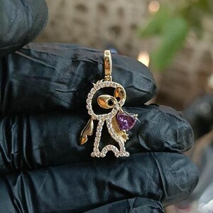 May include: A gold-colored pendant in the shape of a cartoon character, encrusted with small, clear crystals. The character holds a heart-shaped, purple gemstone. The pendant is suspended from a gold-colored bail.