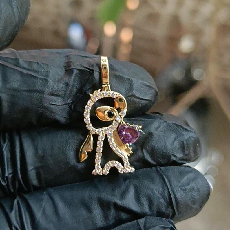 May include: A gold-colored pendant in the shape of a cartoon character, adorned with clear and purple gemstones. The pendant features a heart-shaped purple gemstone and a black gemstone eye. The pendant is suspended from a gold-colored bail.