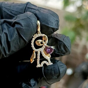 May include: A gold-colored pendant in the shape of a cartoon character, adorned with clear and purple gemstones. The pendant features a heart-shaped purple gemstone and a black gemstone. The character is holding a heart.