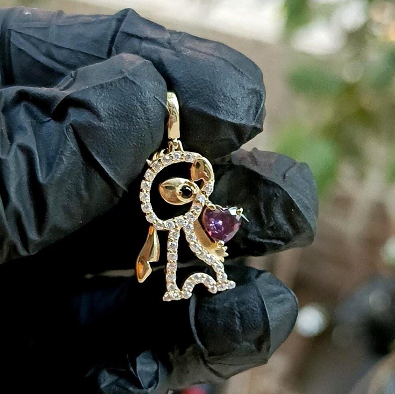 May include: A gold-colored pendant in the shape of a cartoon character, adorned with clear and purple gemstones. The pendant features a heart-shaped purple gemstone and a black gemstone. The character is holding a heart.