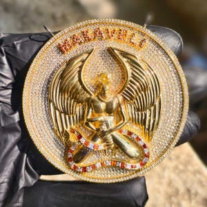 May include: A gold-colored pendant with a detailed design. The pendant features a central figure with wings, surrounded by a ring of sparkling stones. The word "WAKAVILLE" is displayed at the top.