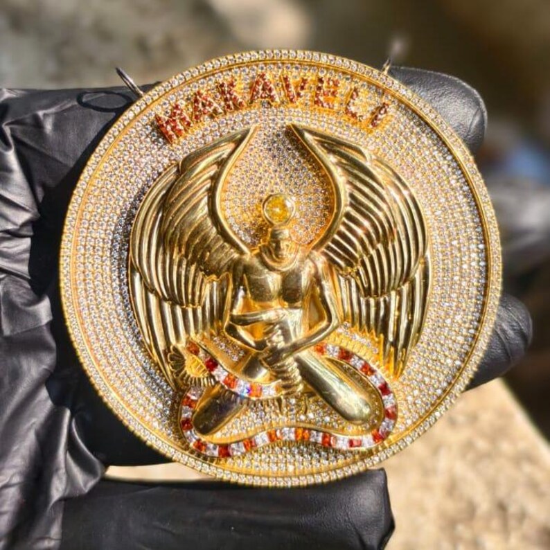 May include: A gold-colored pendant with a detailed design. The pendant features a central figure with wings, surrounded by a ring of sparkling stones. The word "WAKAVILLE" is displayed at the top.