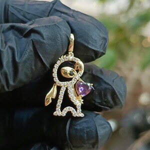 May include: A gold-colored pendant in the shape of a cartoon character, featuring a heart-shaped purple gemstone and small clear gemstones. The pendant has a black gemstone for an eye and is suspended from a matching bail.