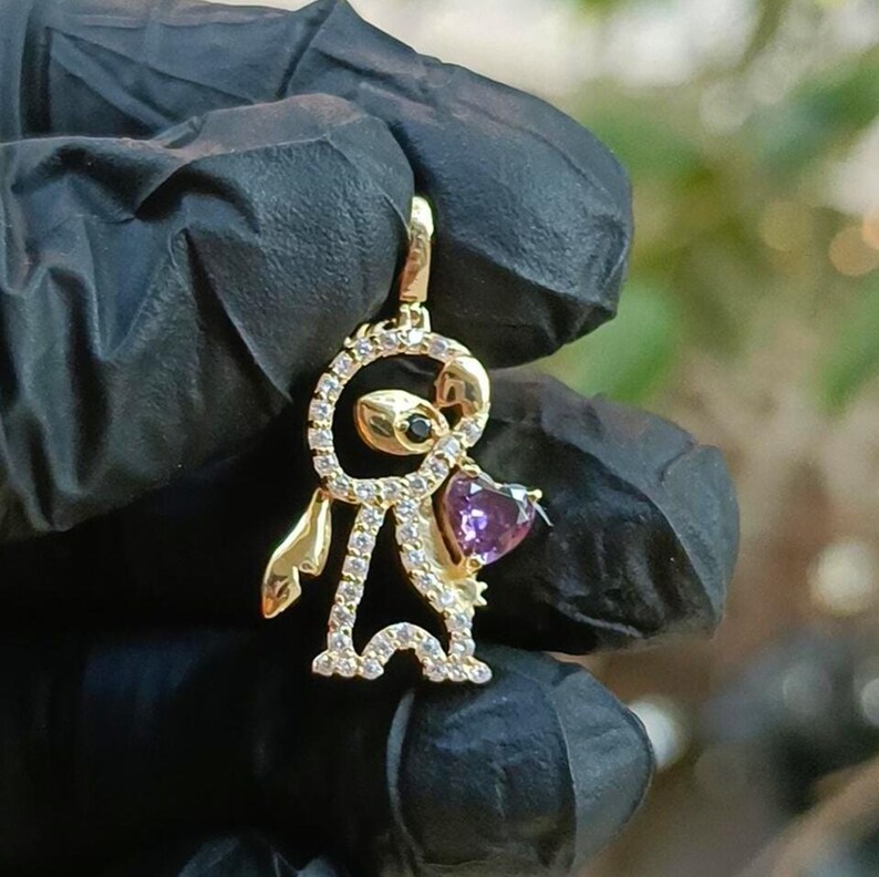 May include: A gold-colored pendant in the shape of a cartoon character, featuring a heart-shaped purple gemstone and small clear gemstones. The pendant has a black gemstone for an eye and is suspended from a matching bail.