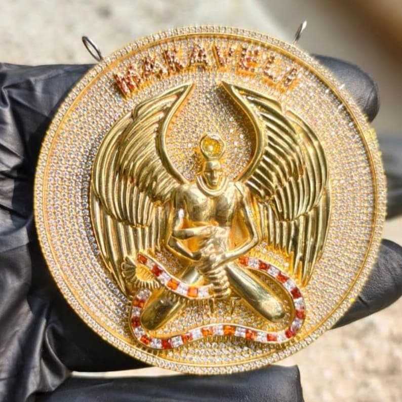 May include: Gold-toned pendant featuring a central figure with outstretched wings, surrounded by a halo of sparkling stones. The pendant has the word "MAKAVELI" in raised letters. A snake-like design with red and white accents wraps around the figure.