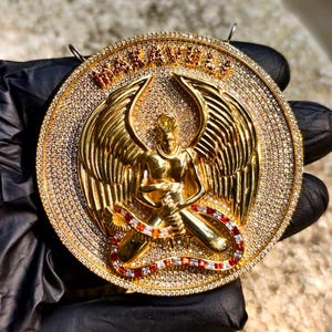 May include: A gold-colored pendant featuring a detailed angel figure with outstretched wings, set within a circular frame. The frame and angel are encrusted with small, sparkling stones. The word "MANAVILLE" is displayed in gold lettering at the top of the pendant.