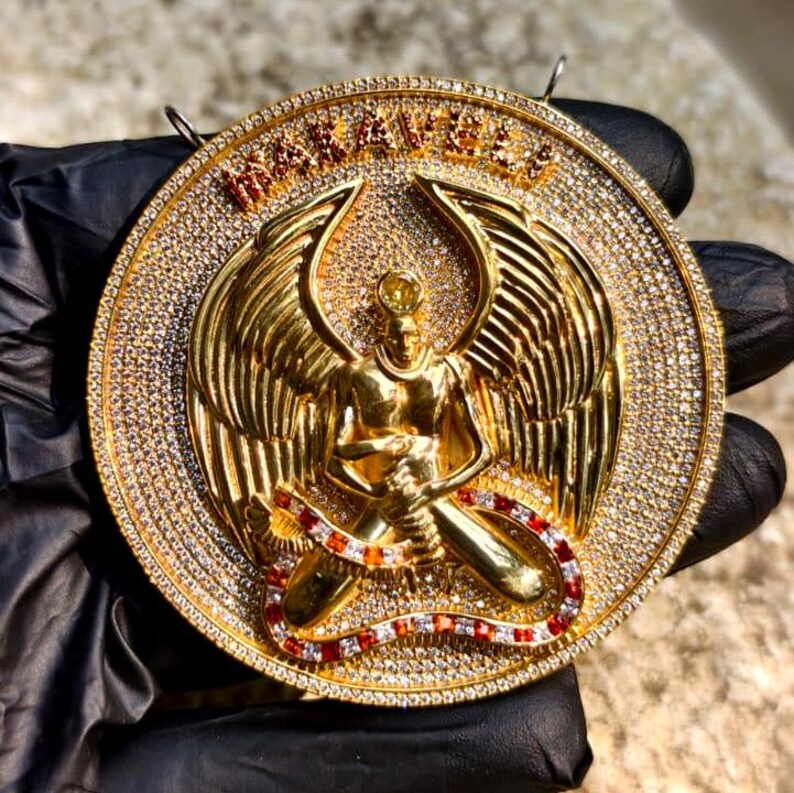 May include: A gold-colored pendant featuring a detailed angel figure with outstretched wings, set within a circular frame. The frame and angel are encrusted with small, sparkling stones. The word "MANAVILLE" is displayed in gold lettering at the top of the pendant.