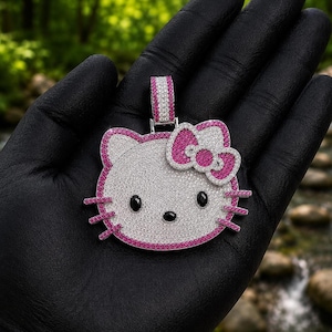 May include: A Hello Kitty pendant with a white face and pink bow and outline. The eyes and nose are black. The pendant is covered in small, sparkling stones. The pendant is held in a black gloved hand.