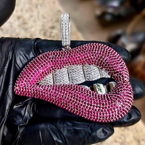 May include: A pendant shaped like a pair of lips, encrusted with pink and clear gemstones. The lips have a silver-colored bale and teeth. The pendant is held by a gloved hand.