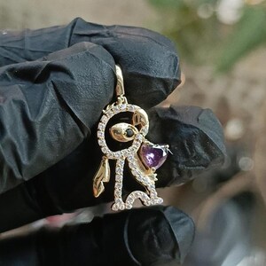 May include: A gold-colored pendant in the shape of a stylized figure, adorned with clear and purple gemstones. The figure has a circular head with a black gem, and a heart-shaped purple gem on the chest. The pendant is held by a gloved hand.