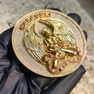 May include: A gold-colored pendant with a detailed angel design. The pendant features a central figure with outstretched wings, surrounded by a halo and encrusted with small, sparkling stones. The word "REMARKABLE" is engraved on the top.