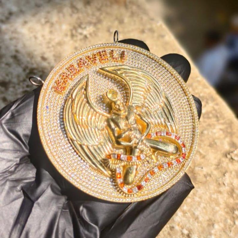 May include: A gold pendant with a detailed design. The pendant features a central figure with wings, surrounded by a ring of sparkling stones. The word "KABARELI" is written across the top in orange letters. The pendant is held by a gloved hand.