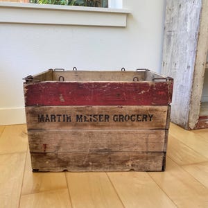 May include: A vintage wooden crate with a red-painted top and the text "MARTIN MEISER GROCERY" in black. The crate has metal hardware and is made of weathered wood, suggesting age and use. It sits on a wooden floor.