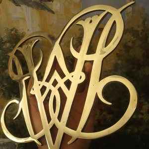 K&ouml;nnte beinhalten: Ein goldfarbenes Metallmonogramm mit einem dekorativen Design. Das Monogramm hat die Form eines stilisierten "W" mit filigranen Details.