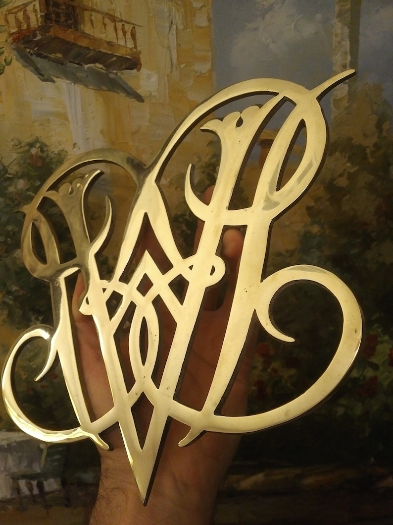 K&ouml;nnte beinhalten: Ein goldfarbenes Metallmonogramm mit einem dekorativen Design. Das Monogramm hat die Form eines stilisierten "W" mit filigranen Details.