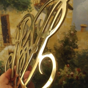 K&ouml;nnte beinhalten: Ein goldfarbenes Metallmonogramm mit einem dekorativen Design. Das Monogramm hat die Form eines stilisierten Buchstabens "P" mit einer Verzierung.