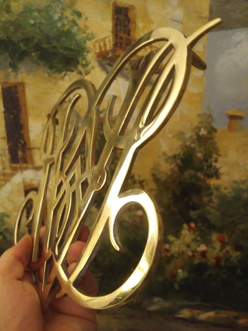 K&ouml;nnte beinhalten: Ein goldfarbenes Metallmonogramm mit einem dekorativen Design. Das Monogramm hat die Form eines stilisierten Buchstabens "P" mit einer Verzierung.