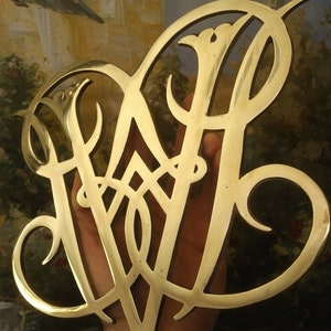K&ouml;nnte beinhalten: Ein goldfarbenes Metallmonogramm mit einem dekorativen Design. Das Monogramm hat die Form eines stilisierten Buchstabens W.