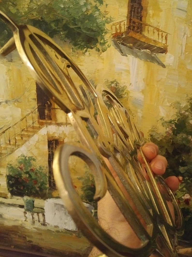 K&ouml;nnte beinhalten: Ein goldfarbener Metall-Topfuntersetzer mit filigranen Designs. Der Untersetzer wird in einer Hand gehalten und befindet sich vor einem Gem&auml;lde eines Geb&auml;udes mit einem Balkon.