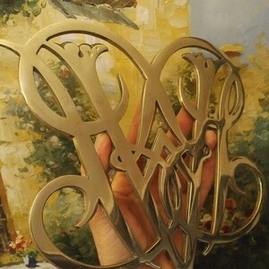 K&ouml;nnte beinhalten: Ein goldfarbenes Metallmonogramm mit einem dekorativen Design. Das Monogramm hat die Form von zwei Buchstaben, m&ouml;glicherweise "W" und "B".