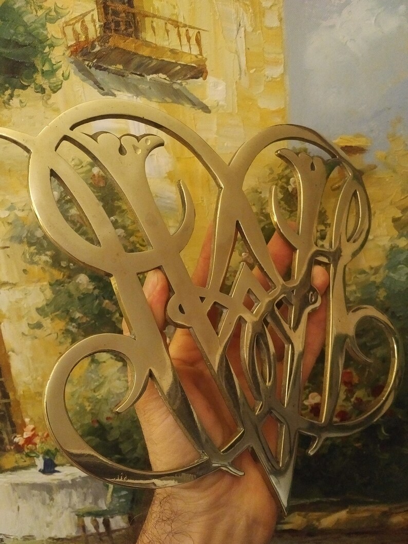 K&ouml;nnte beinhalten: Ein goldfarbenes Metallmonogramm mit einem dekorativen Design. Das Monogramm hat die Form von zwei Buchstaben, m&ouml;glicherweise "W" und "B".