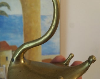 Può includere: Una statuetta di cigno in metallo dorato con un collo lungo e un corpo curvo. Il cigno ha una piccola testa rotonda con un becco e un paio di occhi. Il cigno è seduto su una base piatta con due piccoli piedi.