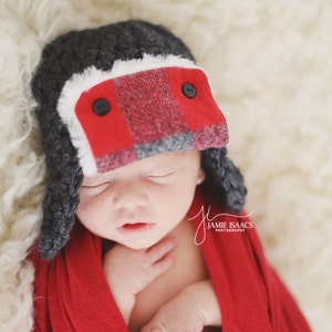 May include: A newborn wearing a gray and red plaid trapper hat with black buttons and a red wrap. The hat has a white faux fur trim. The baby is sleeping on a fluffy, cream-colored surface.