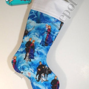 May include: A Christmas stocking featuring characters from the movie "Frozen" on a blue background. The stocking has a white cuff and a loop for hanging. The "Bombshell Depot" logo is in the upper left corner.
