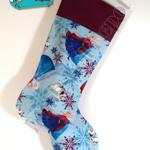 May include: A Christmas stocking featuring a light blue background with images of Elsa and Anna from Frozen, snowflakes, and other characters. The top of the stocking is a deep burgundy color. A black loop is attached for hanging.
