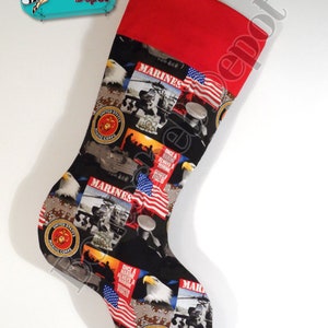 May include: A red and black Christmas stocking with a patriotic theme. The stocking features images of the United States Marine Corps emblem, American flags, eagles, and military vehicles. The text "MARINES" appears on the stocking.