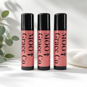 May include: Three cylindrical lip balm tubes standing upright, each featuring a black cap and a coral-pink label. The text MOO + Grace Co is printed vertically in black serif font on the label of each tube, which are arranged in a row.