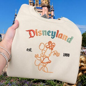 Może przedstawiać: Kremowa bluza z okrągłym dekoltem z napisem Disneyland wyhaftowanym wielokolorową czcionką gradientową. Pod tekstem znajduje się pomarańczowa ilustracja liniowa Myszki Minnie, otoczona czarnym wyhaftowanym napisem est. oraz 1955.