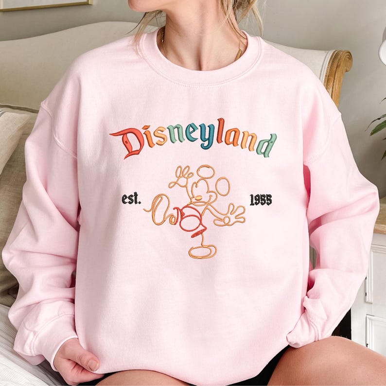 May include: A light pink crewneck sweatshirt featuring the word Disneyland embroidered in a multi-coloured font across the chest. Below the text is an orange outline embroidery of a cartoon mouse character with the text est. 1955 on the sides.