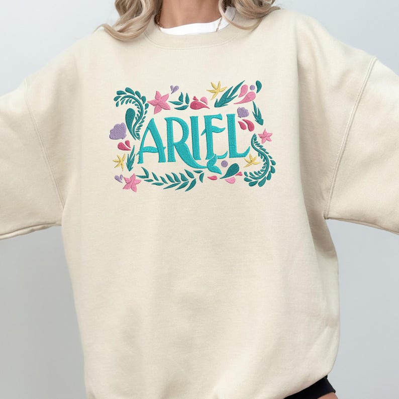 Op de afbeelding: Een cr&egrave;mekleurig sweatshirt met ronde hals en een geborduurd ontwerp op de borst. De tekst ARIEL staat centraal in het groenblauw, omringd door geborduurde botanische en aquatische elementen in tinten groenblauw, roze, geel en paars.