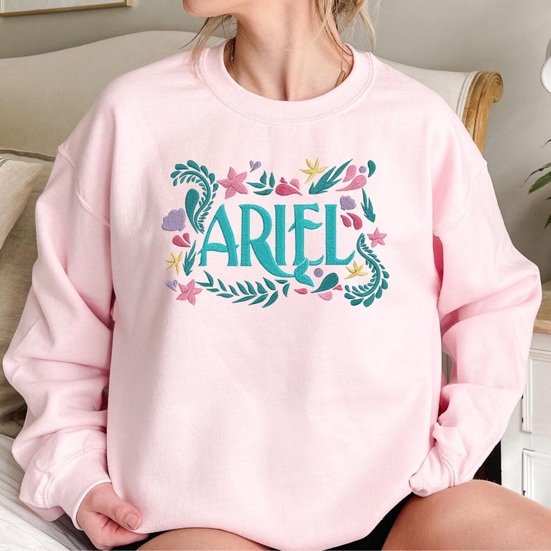 Op de afbeelding: Een lichtroze sweater met ronde hals en een geborduurd ontwerp op de borst. De tekst ARIEL staat centraal in het groenblauw, omringd door kleurrijke geborduurde motieven zoals roze bloemen, groenblauw zeewier, paarse schelpen en gele zeesterren.