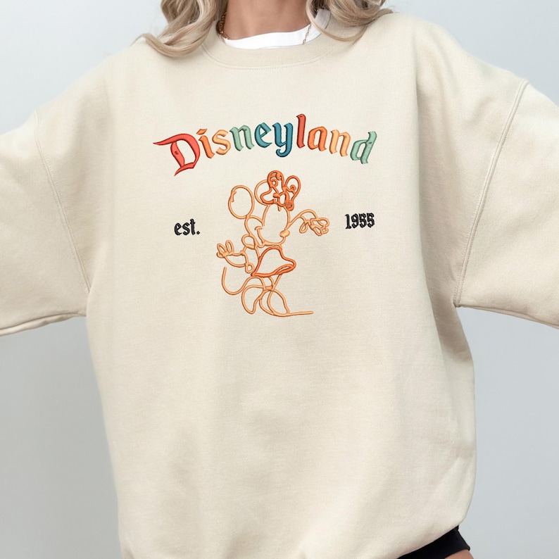 Może przedstawiać: Kremowa bluza z okrągłym dekoltem i wyhaftowanym logo Disneyland w wielokolorowych literach. Pod napisem znajduje się pomarańczowy kontur Myszki Minnie, otoczony czarnym wyhaftowanym napisem est. po lewej stronie i 1955 po prawej.