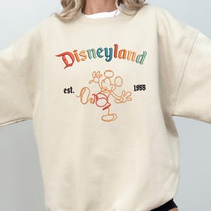 May include: A cream-coloured crewneck sweatshirt featuring an embroidered Disneyland logo in multi-coloured thread. Below the text is an orange line-art illustration of Mickey Mouse, flanked by the black embroidered text est. and 1955.